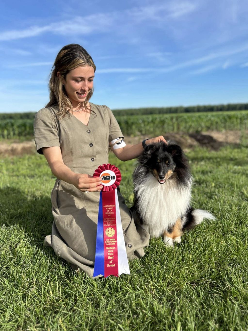 Woman proudly holding a Best of Breed ribbon with her Shetland Sheepdog outdoors.