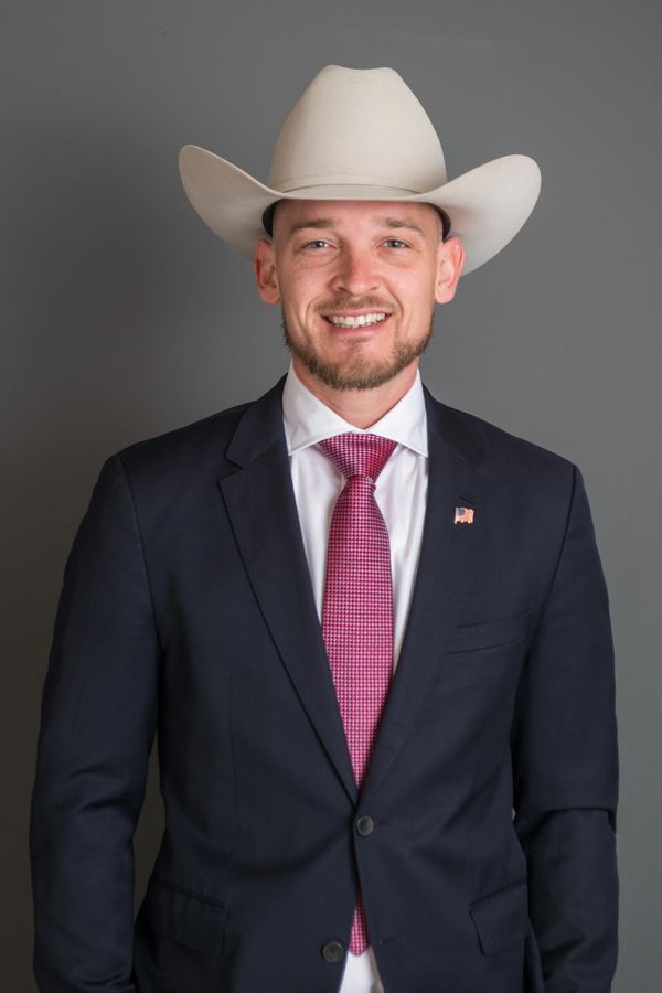 Man in a suit wearing a white cowboy hat and a red tie, smiling.