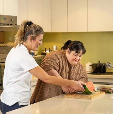 Two women cutting a watermelon together in a kitchen.