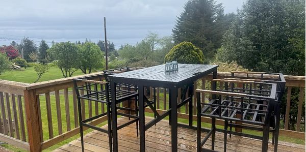 Black metal outdoor table and chairs set on a wooden deck with lush greenery in the background.