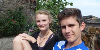 Two young people sitting outdoors near a stone wall and greenery.
