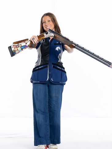 Woman smiling holding a shotgun, wearing shooting vest and jeans.