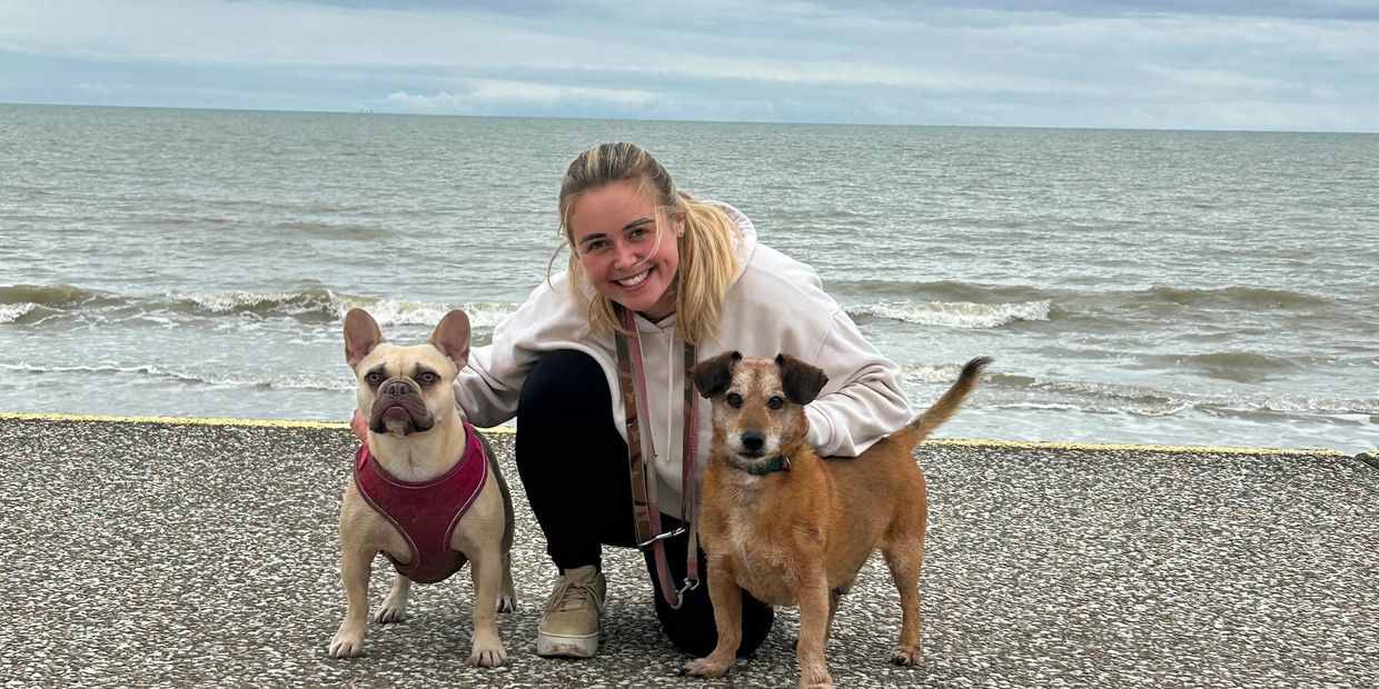 A woman smiles with two dogs by the seaside on a cloudy day.
