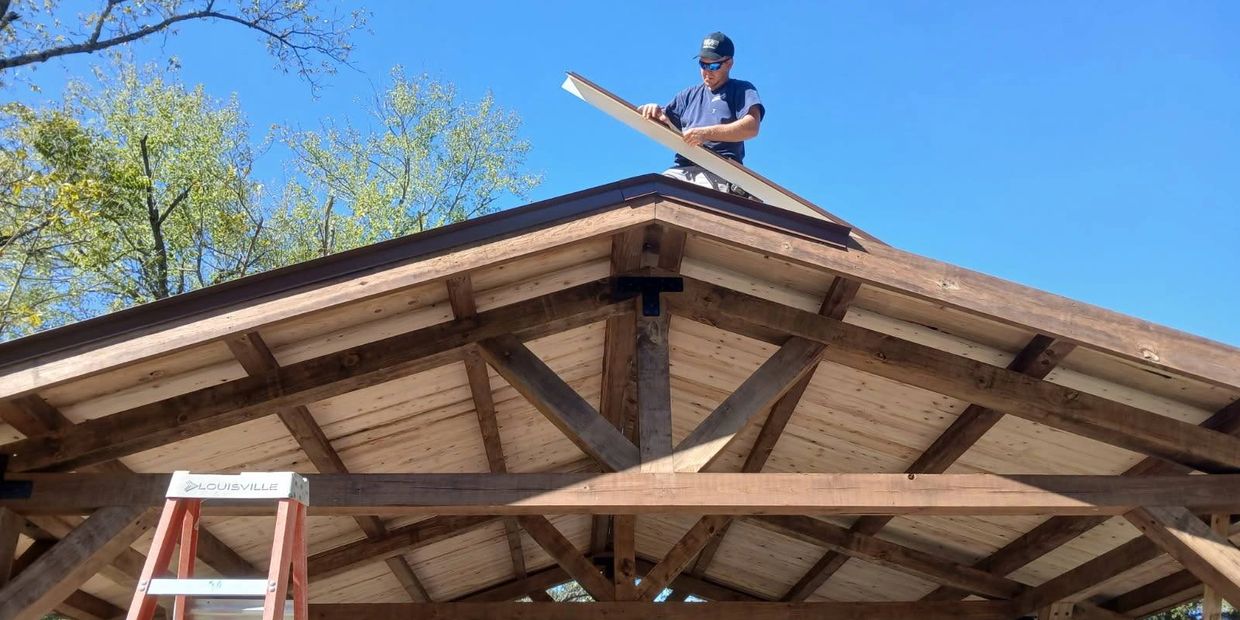 Man working on a wooden roof structure under a clear blue sky.