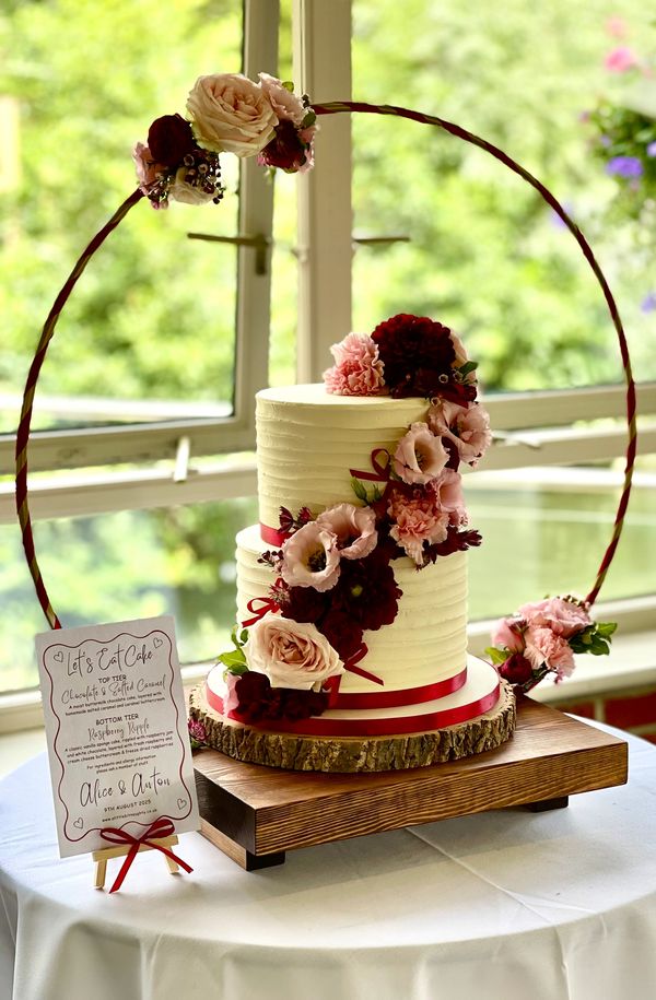 2 tier buttercream cake with pink and red fresh flowers on a wooden stand with gold hoop 