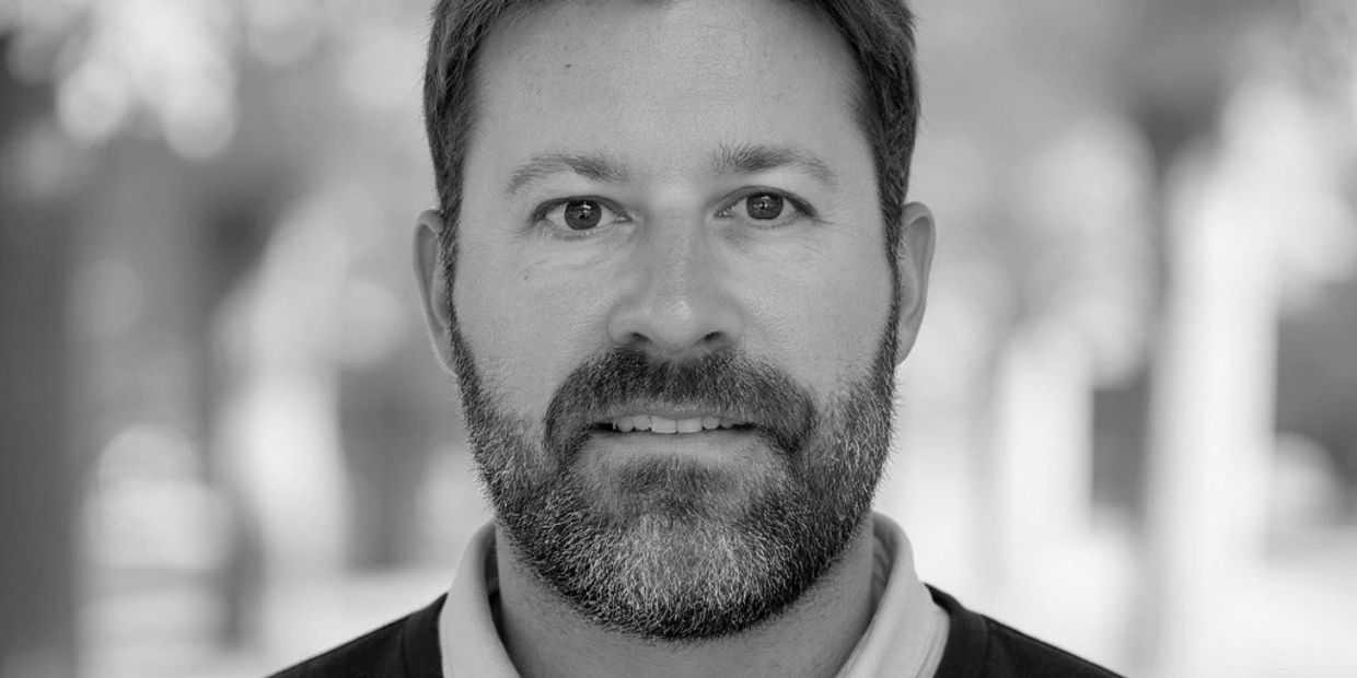 Smiling bearded man outdoors with a blurred green background.