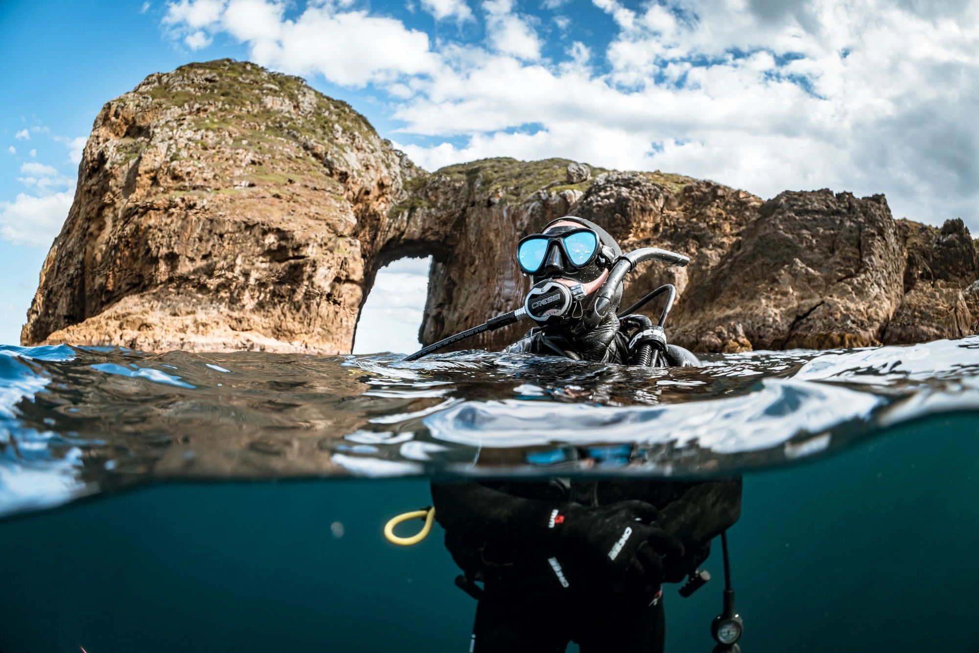 Scuba diver emerging from water near a rocky arch formation.