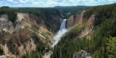 A majestic waterfall cascades into a forested canyon under a blue sky with clouds.