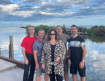 Five adults posing by a waterfront on a partly cloudy day.