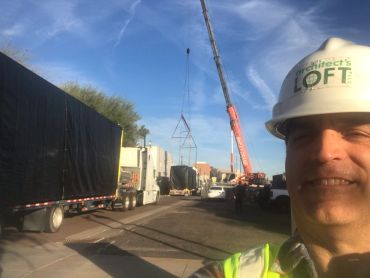 Man in hard hat takes a selfie at a construction site with trucks and a crane.