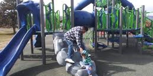 Adult helping child on playground climbing equipment under sunny weather.