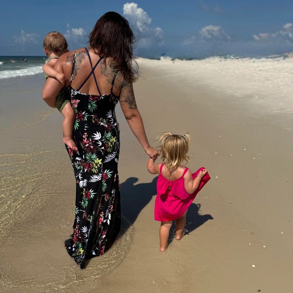 A woman walks on the beach holding hands with a toddler and carrying a baby.
