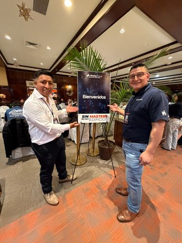 Two men smiling and pointing at a welcome sign at an indoor event.
