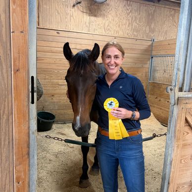 Woman proudly holding a yellow ribbon with her horse in a stable.