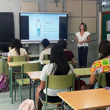 Clase en aula con profesora y estudiantes mirando pantalla.