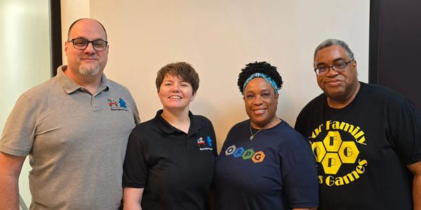 Four adults smiling and posing together indoors, wearing casual shirts with game-related logos.