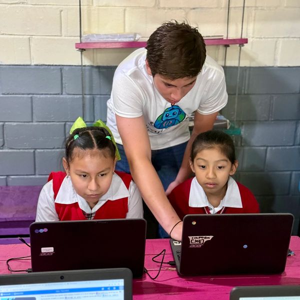 Teacher assists two girls in school uniforms using laptops for programming.