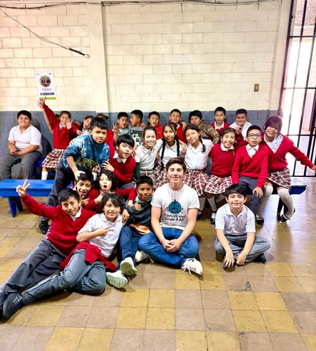 A lively group of schoolchildren and a young adult sitting and posing in a classroom.