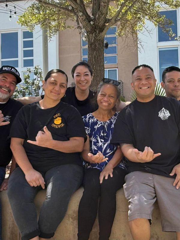 Group of six people smiling and making shaka hand signs outdoors.