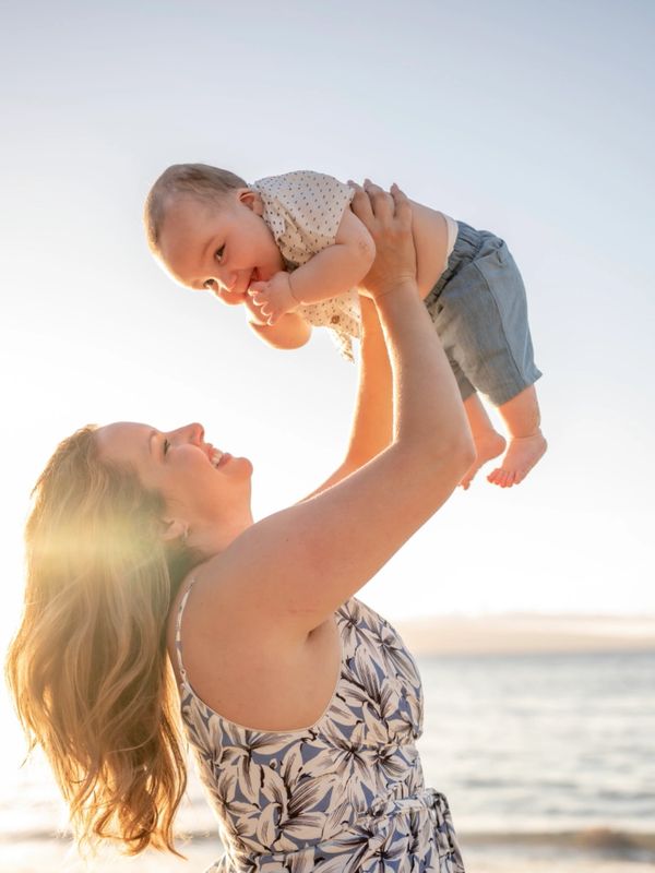 Mother joyfully lifts her smiling baby by the beach at sunset.
