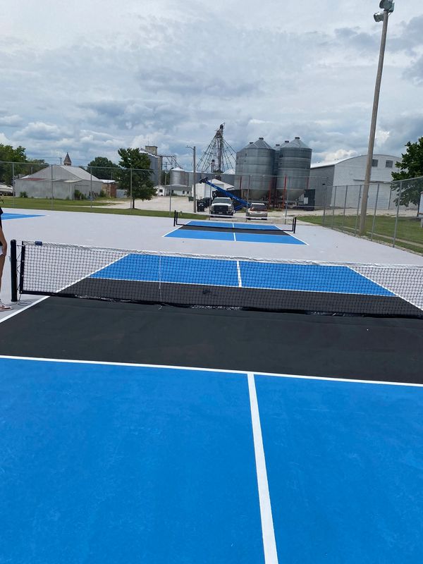 Two pickleball courts with blue and black surfaces under a cloudy sky.
