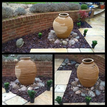 A decorative ribbed clay pot surrounded by rocks and small plants in a garden corner.