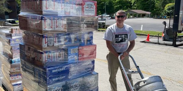 man standing beside pallet of beer taller than he is, wheeling a keg on a dolly