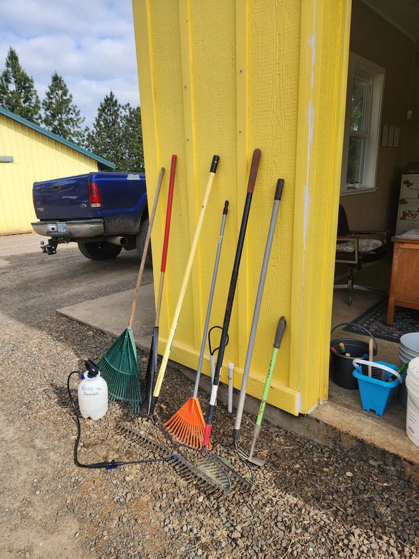 Garden tools and a sprayer leaning against a yellow shed on a gravel surface.