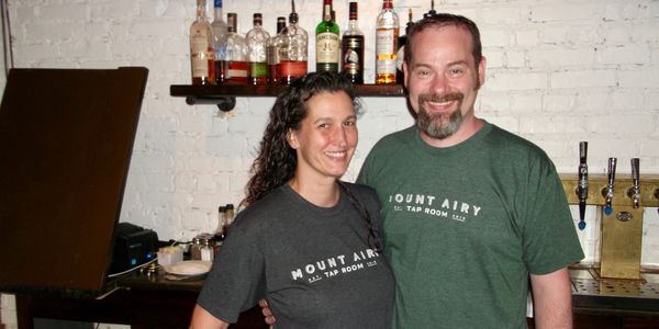 Two people smiling at a bar wearing Mount Airy Tap Room shirts.
