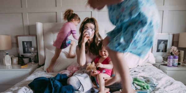 A tired mother drinking coffee while kids play energetically on the bed.