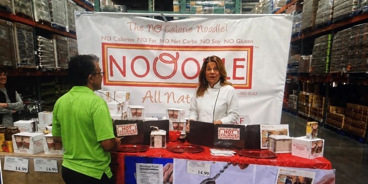 Woman in white coat promoting NoOodle noodles at a store booth.