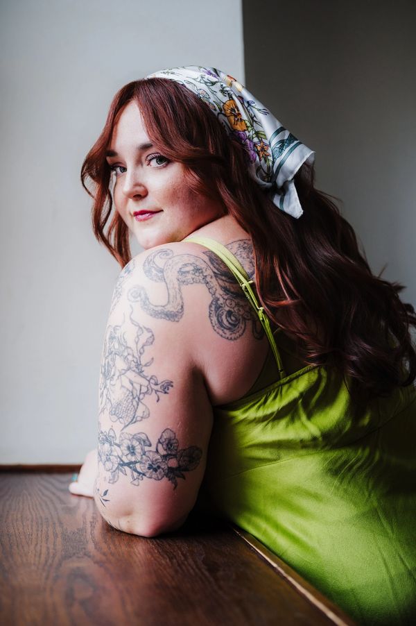 Woman with red hair and floral scarf showcasing detailed tattoos on her arm and back.
