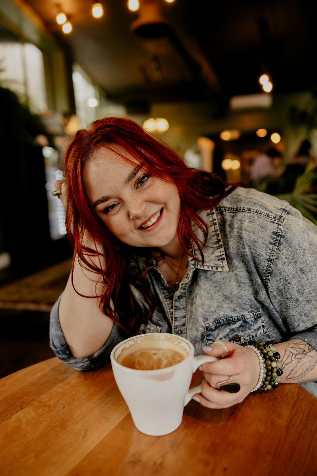 A smiling red-haired woman enjoys coffee at a cozy café.