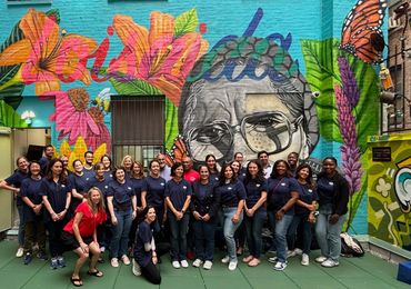 Group of people posing in front of a colorful mural with flowers and a face.