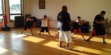 A martial arts instructor teaches a group of children punching techniques in a sunlit gym.