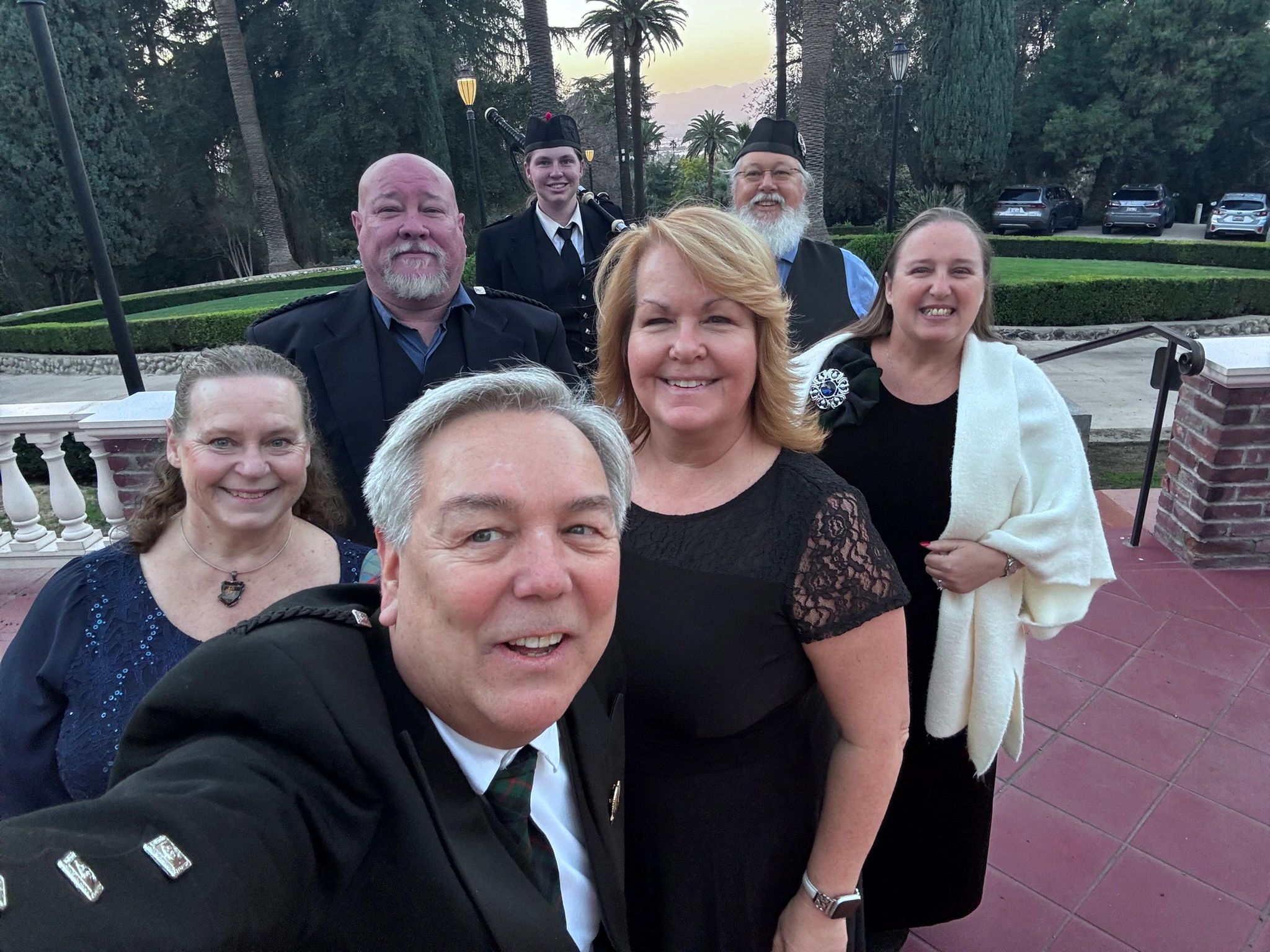 A group selfie of seven people dressed in formal and traditional Scottish attire outdoors.