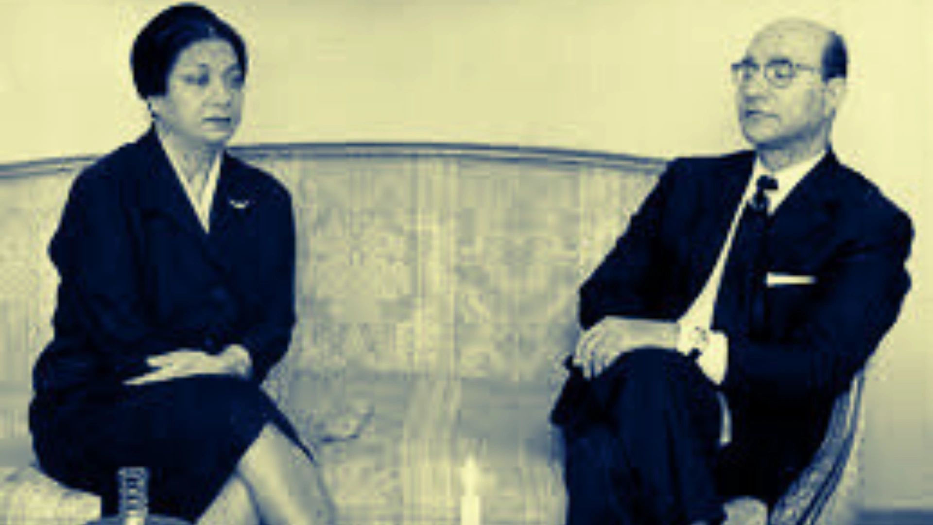 Two individuals in formal attire seated on a couch, engaged in conversation.