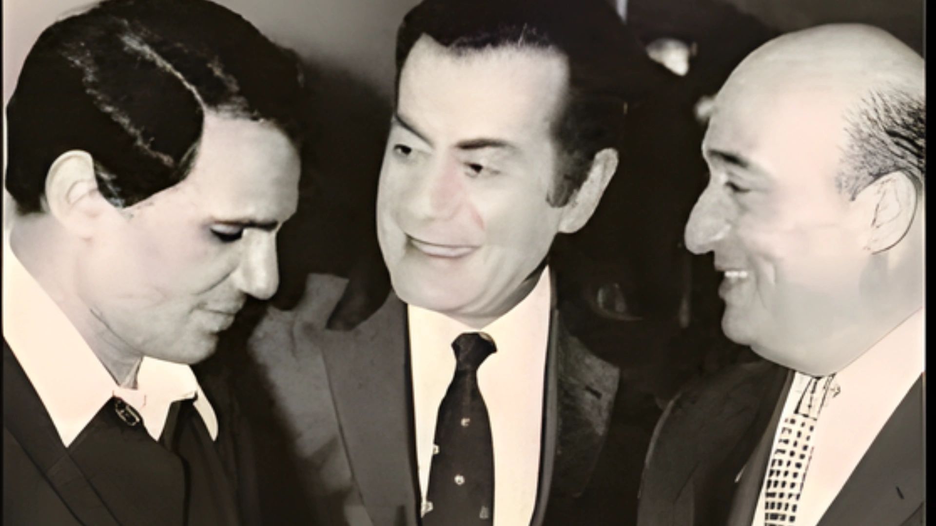 Three men in suits share a moment, with one smiling warmly at the others.