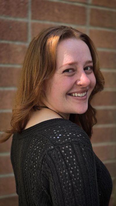 Smiling woman with red hair in a black sweater against a brick wall.