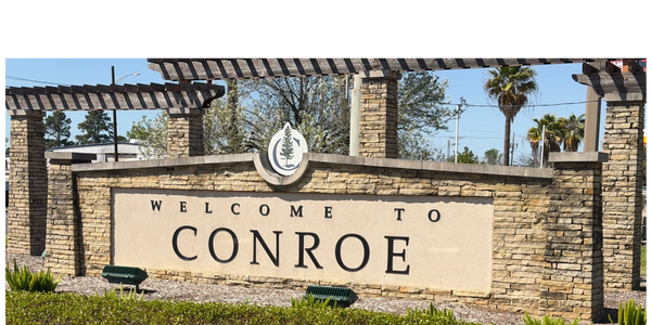 Stone sign welcoming visitors to Conroe with greenery and palm trees nearby.