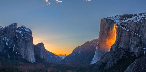 Sunset lights up a waterfall in a mountain valley with snow-capped peaks.