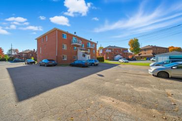 A sunlit residential parking lot with brick apartment buildings and several parked cars.