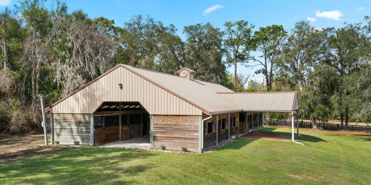 Large wooden barn with metal roof in a grassy area surrounded by trees.