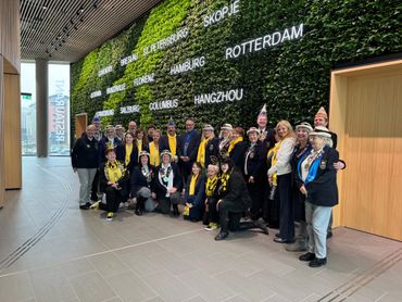 Group of people in colorful scarves and hats posing indoors by a green wall with city names.