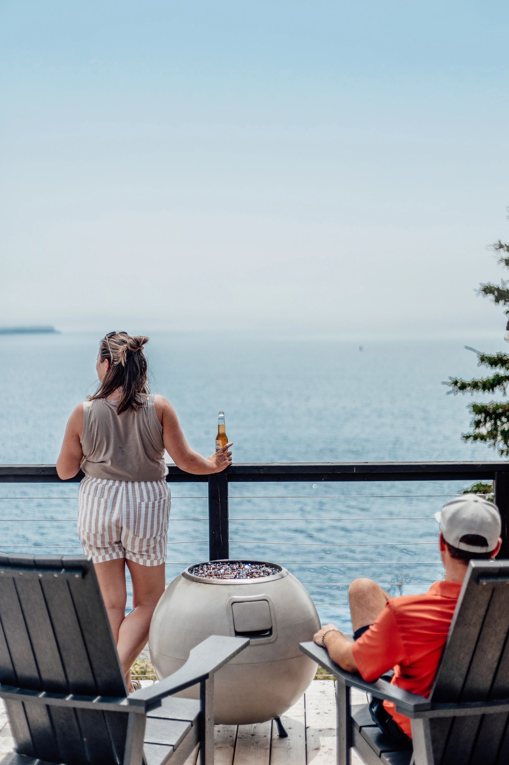 Woman overlooks ocean from private deck at Nova Scotia glamping dome with man sitting nearby.
