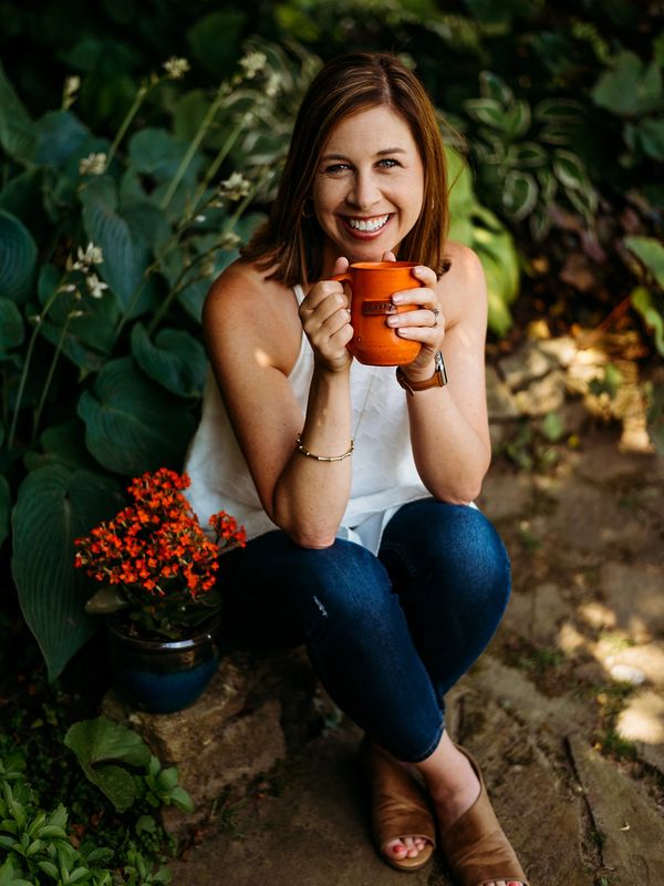 White woman with brown hair, sitting outide next to flowers.  Holding an orange coffee mug