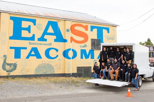 A group of people posing in and around a truck in front of an "EAST TACOMA" mural.
