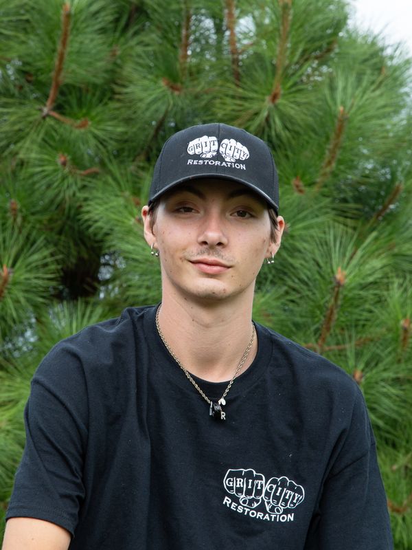 Young man wearing a black "Grit City Restoration" cap and shirt, posing outdoors.