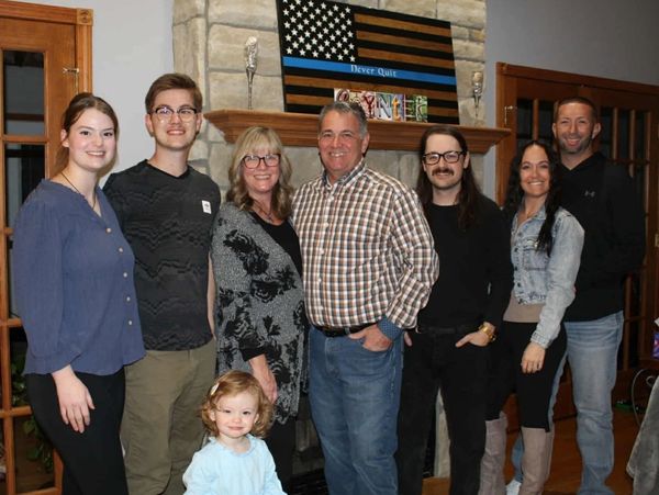 A smiling family posing indoors in front of a stone fireplace with a patriotic flag.