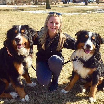 Young blonde haired woman smiling at a park with two Bernese Mountain Dogs on each side of her